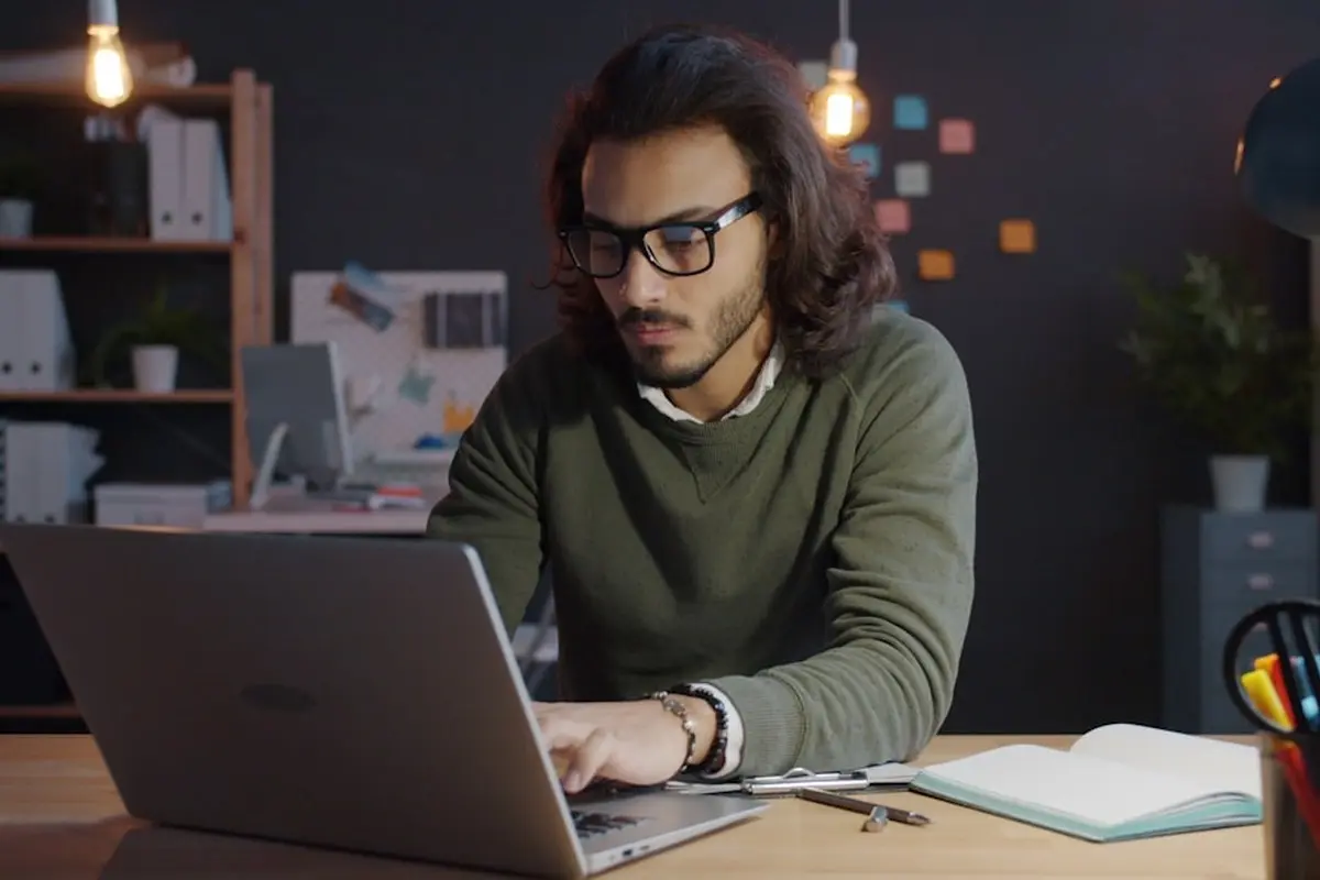 man standing and facing whiteboard with writings near woman pointing at it and facing man sitting beside table with laptop indoors
