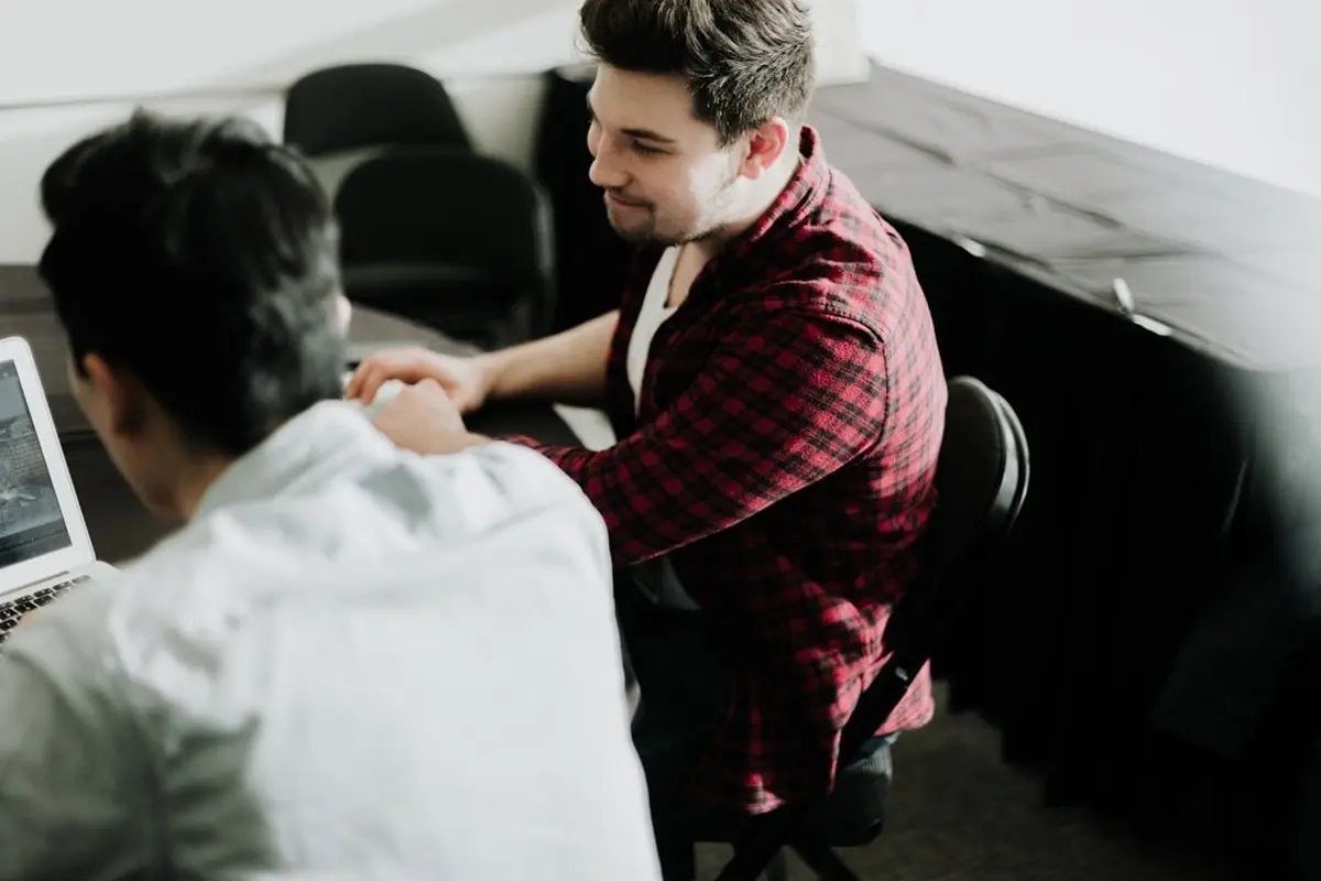 man standing and facing whiteboard with writings near woman pointing at it and facing man sitting beside table with laptop indoors